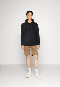 Black hoodie with "GAP" logo, paired with khaki shorts. Model wears white socks and sneakers. Simple, casual clothing design.