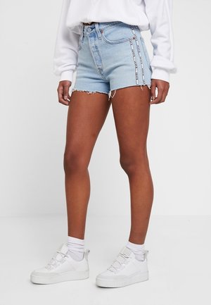 Personne portant un short en denim bleu clair avec des bandes blanches à logo sur les côtés, un haut blanc à manches longues, des chaussettes blanches et des baskets blanches.