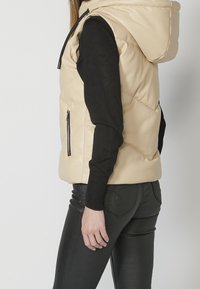 Personne portant un gilet sans manches matelassé beige à capuche sur un haut noir à manches longues et un pantalon noir ajusté avec des poches arrière détaillées.