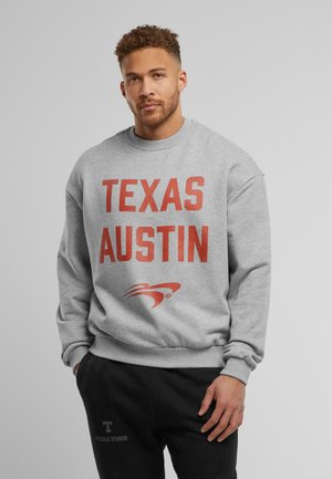 Grå sweatshirt med stor rød tekst "TEXAS AUSTIN" på fronten, med et logo nedenunder. Ribbet afslutning ved ærmerne og taljen, blød stoftekstur.