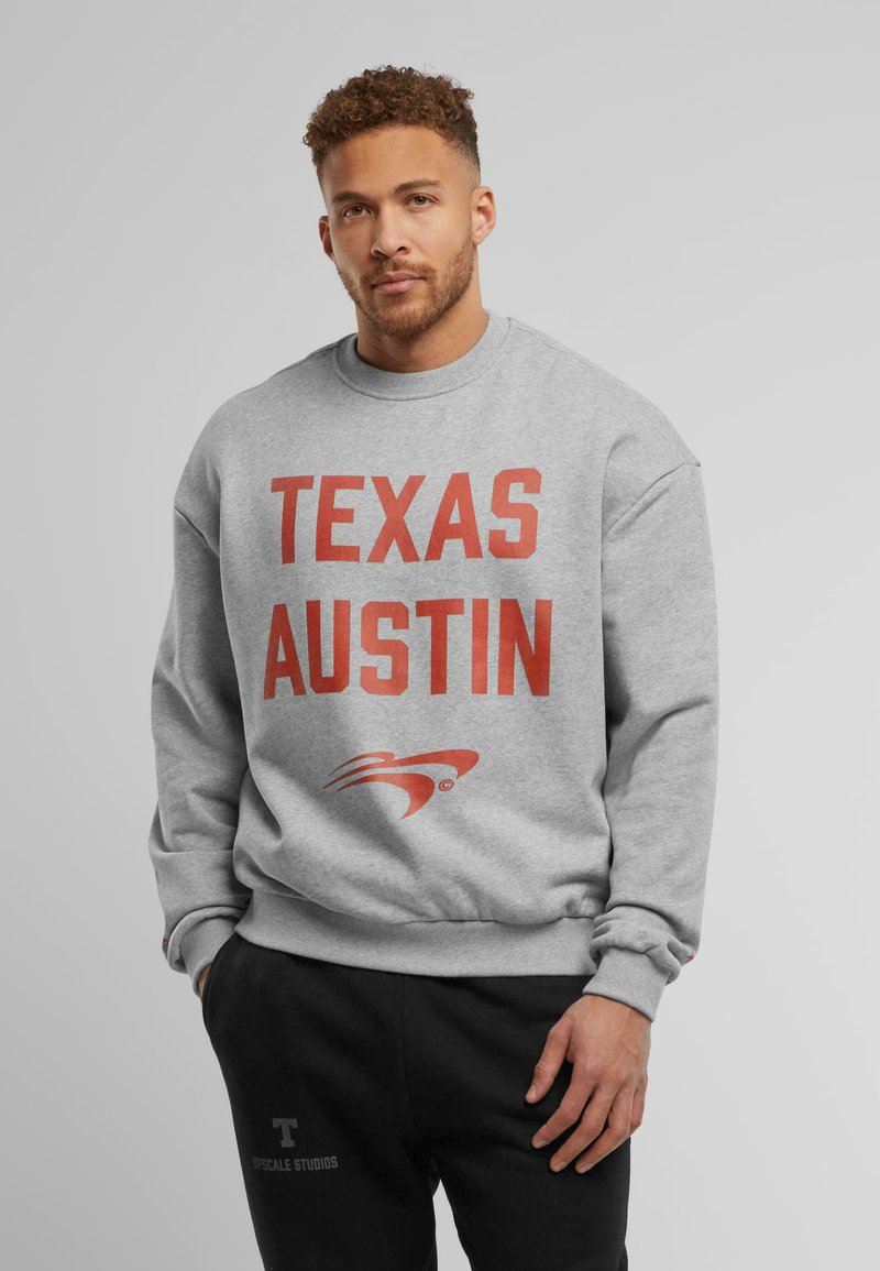 Sweatshirt gris avec un grand texte rouge "TEXAS AUSTIN" à l'avant, accompagné d'un logo en dessous. Manches et taille côtelées, texture de tissu douce.