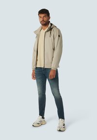 Lichtgrijze hoodie-jack met een ritssluiting aan de voorkant, elastische manchetten en een losse pasvorm, gecombineerd met een beige t-shirt en blauwe jeans. Witte sneakers.