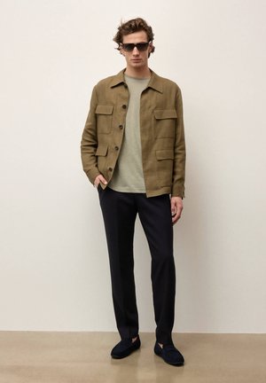 Homme portant des lunettes de soleil, veste boutonnée vert olive sur une chemise vert clair, pantalon noir et mocassins foncés, debout contre un mur beige.