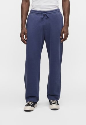 Homme portant un pantalon de survêtement bleu marine à cordon et des baskets basses noires avec semelles et lacets blancs, debout sur un fond blanc.