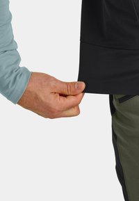 Schwarzes Shirt mit glatter Textur, leicht verlängertem Saum und langen, hellblauen Ärmeln. Eine Hand hält den unteren Rand des Shirts.