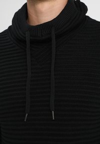 Zwarte gebreide trui met een hoge, ribbelige kraag en trekkoorden. De textuur is ribbelig en het ontwerp heeft een relaxte pasvorm.