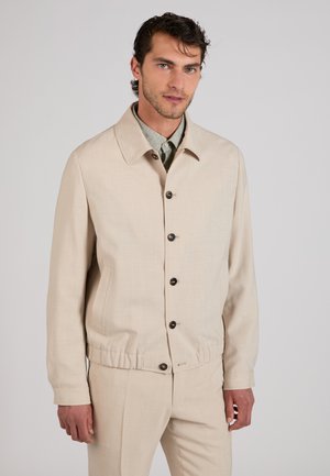 Homme portant une veste beige boutonnée avec taille élastique et pantalon assorti, debout sur un fond uni gris clair.