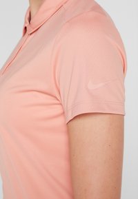 Světle růžové polokošile s měkkou texturou, s límečkem a krátkými rukávy. Na levém rukávu je jemné logo Nike.