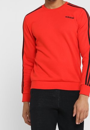 Mężczyzna w czerwonej bluzie Adidas z czarnymi paskami na rękawach i czarnych spodniach, stojący na jednolitym tle.