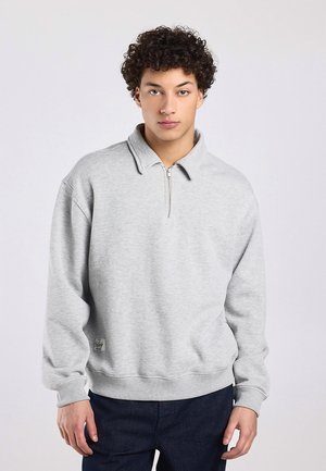 Jeune homme aux cheveux bouclés portant un pull zippé gris clair et un pantalon foncé, debout devant un fond blanc uni.