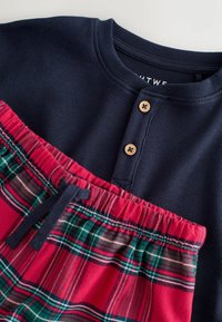 Marineblaue gerippte Bluse mit zwei Holzknöpfen, kombiniert mit rot karierten Shorts mit grünen und weißen Akzenten sowie einem elastischen Bund.