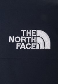 Černá látka s vyšívaným bílým logem obsahujícím text "THE NORTH FACE" a charakteristický půlkruhový motiv.