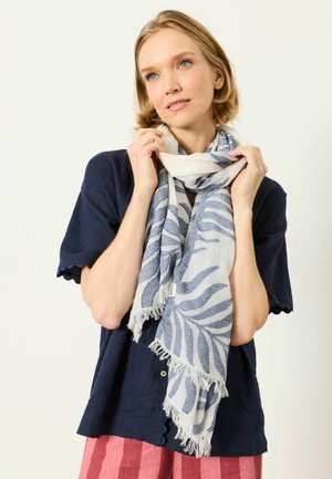 Moteris su trumpais blondiniais plaukais, dėvinti tamsiai mėlyną marškinį, rožinius dryžuotus kelnes ir baltai mėlyną raštuotą šaliką, laikanti šaliką prie veido.