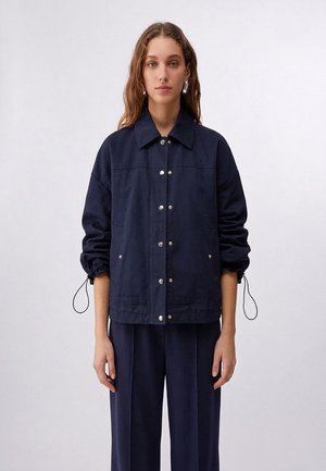 Donna con capelli lunghi e mossi indossa una giacca blu navy con bottoni, polsini elasticizzati e pantaloni abbinati a gamba larga, in piedi davanti a uno sfondo uniforme.