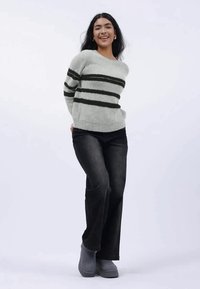 Lys grå genser med svarte striper, myk tekstur, kombinert med mørke jeans og grå støvler. Modellen poserer med hendene på hoftene foran en ensfarget bakgrunn.