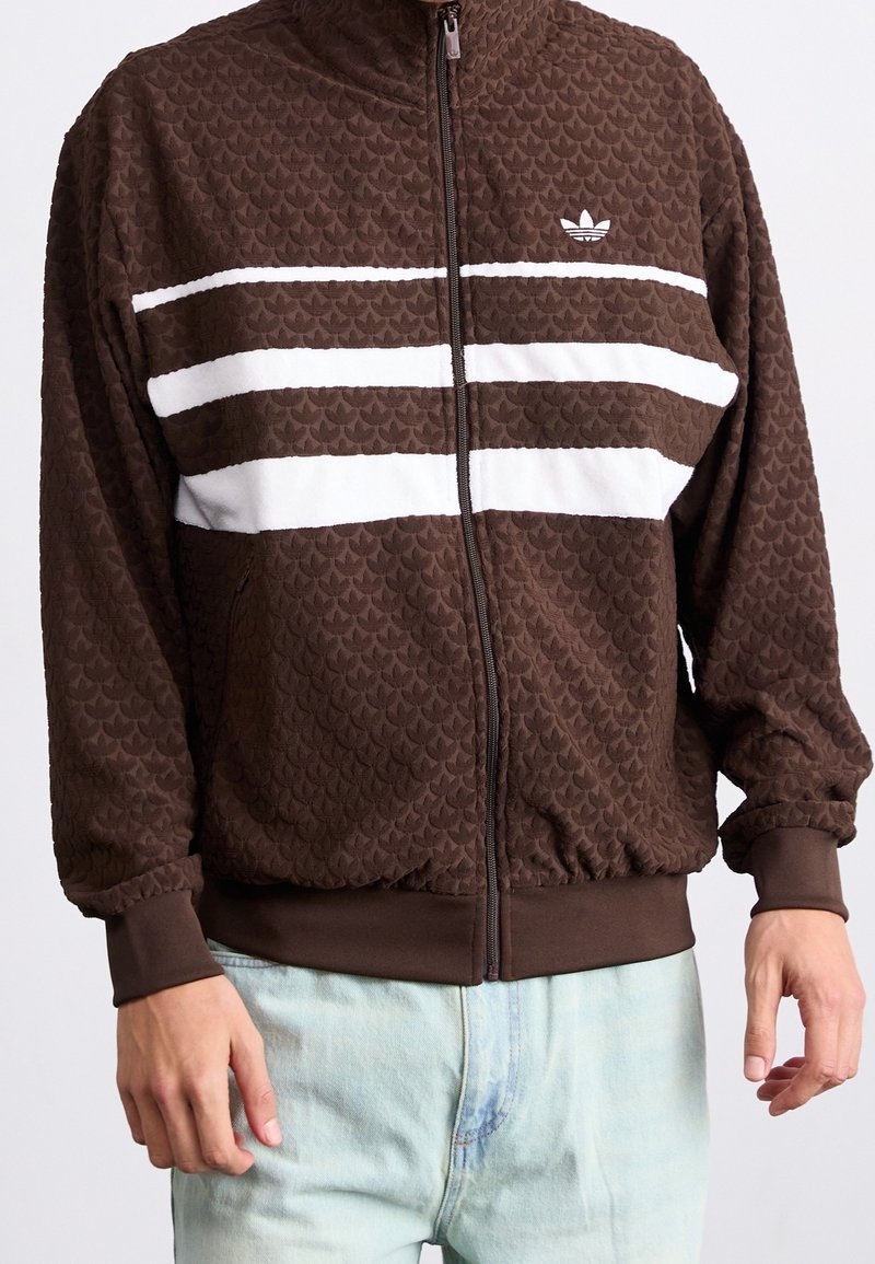 Ruskea vetoketjullinen takki, jossa on teksturoitu kuvio, ja rinnan kohdalla valkoiset vaakaraitat sekä logo vasemmalla puolella.