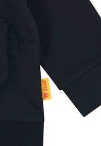 Zwarte sweatshirt met een gerimpelde zak, ribgebreide manchetten en een gele label met rode tekst aan de onderrand. Gladde stoftextuur.