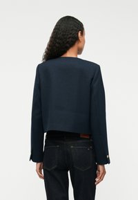 Marineblå kort jakke med teksturert stoff, rett snitt, enkel rund hals og lange ermer. Kombinert med mørke denimjeans.