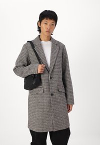 Abrigo a cuadros en tonos marrón y crema con solapas muesca, dos bolsillos frontales y botones. Combinado con un pequeño bolso negro de hombro.