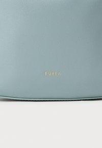 Bolsa de couro azul-claro com uma superfície texturizada. Apresenta um logo dourado "FURLA" na parte inferior, design minimalista com bordas arredondadas.