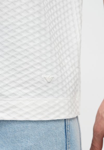 Λευκό υφασμάτινο t-shirt με διαμάντι μοτίβο, με μικρό λευκό λογότυπο στο τελείωμα, συνδυασμένο με ανοιχτό μπλε τζιν.