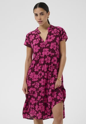 Nő, aki lila, térdig érő ruhát visel rózsaszín virágmintával, rövid ujjú, V-nyakkivágással, és arany karika fülbevalóval, valamint karkötőkkel.