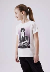 T-shirt en coton blanc avec un imprimé graphique d'une fille aux cheveux noirs tressés, portant une robe à pois, et le mot "MERCREDI."