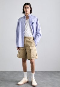 Camisa de botones a rayas en azul claro y blanco, pantalones cortos tipo cargo en beige, calcetines blancos y zapatillas de crema con detalles marrones. Conjunto simple y casual.