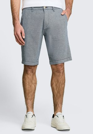 Mann trägt hellgraue, maßgeschneiderte Shorts, weiße Turnschuhe und ein weißes Hemd, mit einer Hand in der Tasche, steht mit leicht gespreizten Beinen.