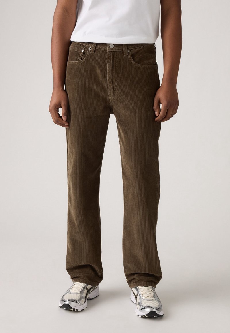 Homme portant un pantalon droit en velours côtelé marron, un T-shirt blanc et des baskets blanches et grises, debout devant un fond uni.