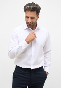 Camicetta bianca con colletto button-down, maniche lunghe e polsini texturizzati, abbinata a pantaloni blu navy. Tessuto liscio e vestibilità su misura.