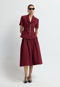 Donna che indossa un blazer corto bordeaux con maniche a breve e bottoni, una gonna midi abbinata, décolleté nere con cinturino a T, occhiali da sole a occhio di gatto e orecchini pendenti dorati.