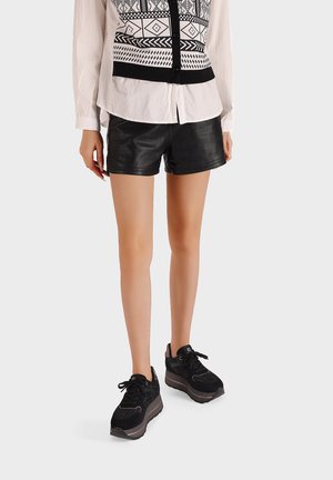 Pantaloni corti in pelle nera abbinati a una camicia bianca con bottoni decorata con un motivo geometrico nero, indossati con scarpe da ginnastica nere.