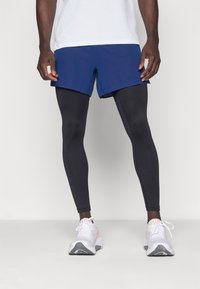 Korte marineblauwe shorts over zwarte leggings, gecombineerd met witte sportschoenen met grijze accenten en roze details.