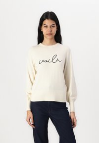 Pull en tricot crème à manches longues et ourlet côtelé. Présente un texte en cursive noir "voilà" sur le devant. Texture lisse. Associé à un jean foncé.