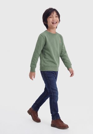 Garçon souriant marchant, portant un pull vert, un jean bleu foncé et des bottes marron à lacets sur un fond clair uni.