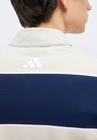 Άτομο που βρίσκεται με την πλάτη και φοράει μια μπεζ και ναυτική μπλούζα με ένα μικρό λευκό λογότυπο Adidas στον άνω πίσω γιακά.