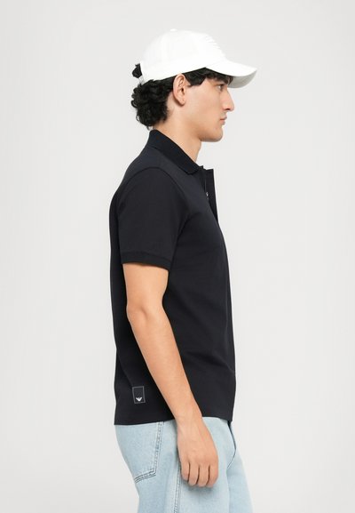 Man som bär en vit baseballkeps, svart kortärmad polo-shirt och ljusblå jeans, står i profil mot en enfärgad bakgrund.