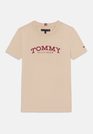 T-shirt beige a maniche corte in cotone con la scritta "TOMMY HILFIGER" in lettere rosse sul petto e piccolo logo a forma di bandiera sulla manica sinistra.