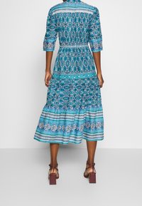 Abito maxi blu con fantasia, maniche a tre quarti, vita aderente e gonna a strati. Presenta disegni intricati e dettagli decorativi.