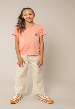 Fille souriante avec une longue queue de cheval, portant un t-shirt pêche avec un détail en forme de cœur, un pantalon large crème et des baskets orange et blanches, debout de manière détendue.
