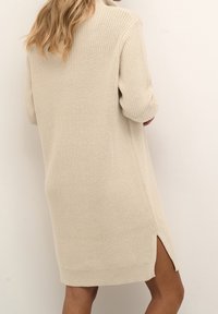Femme portant une robe pull en tricot crème, longueur genou, avec manches longues et fente latérale, vue de dos.