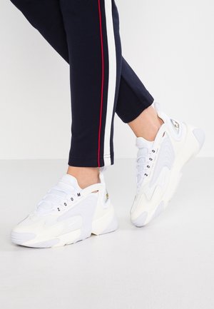 Personne portant des baskets blanches de sport avec le texte « ZOOM », associées à un pantalon noir avec des bandes verticales rouges et blanches.
