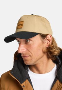 Casquette beige avec un bord foncé, arborant un écusson en cuir carré. La casquette est fabriquée en tissu durable et est portée avec une veste marron.