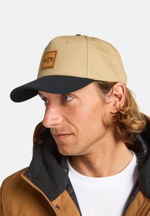 Beige cap with a dark brim, featuring a square leather patch. The cap is made of durable fabric, and worn with a brown jacket.