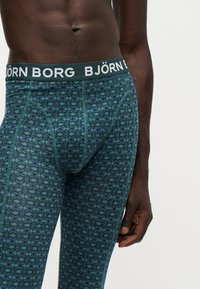 Buste masculin portant un sous-vêtement long ajusté bleu et vert à motifs avec une ceinture de marque Björn Borg sur fond blanc.