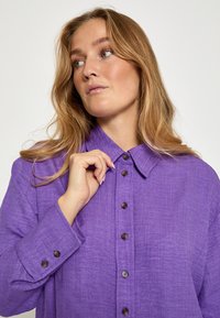 Lilla linned skjorte med knaplukning foran, spidse kraver og lange ærmer. Har brune knapper og en tekstureret stof.