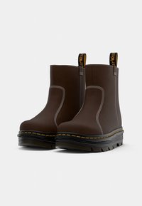Botas de tobillo marrones con un acabado suave, costuras blancas, una suela negra gruesa y acentos amarillos en el borde y en la tira de agarre.