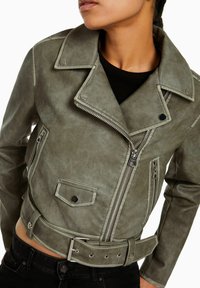 Zkrácená zelená kožená motorkářská bunda se stříbrnými zipy, patentkami a páskem v pase. Má strukturovaný materiál a několik kapes.