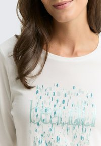 Close-up van een vrouw die een wit shirt draagt met turquoise abstracte patronen en het woord "mooi" gedrukt op de voorkant, die zachtjes glimlacht.